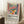 May include: Colorful Matisse-inspired still life poster in light wood frame leaning against white wall. Features vibrant orange vase with red flowers, green leaves, blue teapot, green pitcher, and fruit on pink table with striped background. Displayed in cozy room setting with white coffee cup on wooden stool and beige textured rug.