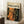 May include: Framed art poster leaning against white wall corner featuring abstract expressionist painting with vibrant orange, dark green, and black paint splatters and drips. Wooden frame, cream matting with "CHAOS AND CREATIVITY POLLOCK 1912-1956" text. Small wooden side table with white coffee cup and saucer nearby, beige textured rug below.