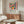 May include: Colorful Matisse-style poster in wooden frame hanging above walnut credenza in modern living room. Artwork features vibrant still life with flowers in orange vase, fruits, and pottery in bold reds, blues, yellows, and greens. Room has cream walls, white curved chair, glass oval coffee table, and brass floor lamp. Natural lighting creates warm, contemporary atmosphere.
