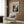 May include: Cream-colored poster featuring Jackson Pollock's abstract expressionist drip painting in black and gray tones, displayed in light wooden frame on wood-paneled wall. Modern reading nook with beige upholstered chair, geometric patterned pillow, brass floor lamp, and dark round side table with open book. Natural lighting from nearby window with sheer curtains.