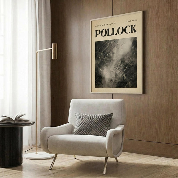 May include: Cream-colored poster featuring Jackson Pollock's abstract expressionist drip painting in black and gray tones, displayed in light wooden frame on wood-paneled wall. Modern reading nook with beige upholstered chair, geometric patterned pillow, brass floor lamp, and dark round side table with open book. Natural lighting from nearby window with sheer curtains.