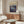 May include: Abstract art poster with swirling blue, purple and teal paint patterns resembling cosmic or fluid forms, displayed in thin wooden frame above mid-century modern walnut credenza. Room features white curved chair, glass oval coffee table, white table lamp, and neutral walls with natural lighting.