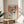 May include: Colorful Matisse-inspired still life poster in wooden frame on round wood table next to beige chair and plant. Artwork shows orange vase with red flowers, green leaves, blue pitcher, and yellow-red fruits on pink surface with striped background in vibrant fauve style colors.