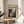 May include: Framed Jackson Pollock exhibition poster in light wood frame, featuring abstract black and white drip painting with "CHAOS AND CREATIVITY 1912-1956 POLLOCK" text, displayed on round wooden side table next to beige upholstered chair with plant shadows from window light on cream wall