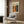 May include: Modern living room scene with beige armchair, geometric pillow, brass floor lamp, and dark round side table with open book. Wood-paneled wall displays framed art poster featuring abstract expressionist painting with orange, black, and green paint splatters and drips. Natural light from window with white curtains illuminates the contemporary interior space.