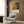 May include: Jackson Pollock exhibition poster with abstract yellow and green paint drip artwork mounted on wood-paneled wall above beige upholstered armchair with geometric patterned pillow. Modern reading corner features gold floor lamp and dark round side table with open book. Warm neutral interior with wood grain texture and natural lighting from window with white curtains.