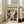 May include: Large framed abstract art print leaning against white wall next to beige upholstered chair in bright living room. Poster features bold black and white paint splatters and drips in energetic composition with "POLLOCK" text at top and "CHAOS AND CREATIVITY 1912-1956" subtitle. Natural wood frame, aged beige background texture, sunlight streaming through windows onto hardwood floors.