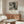 May include: Black and white abstract expressionist artwork hanging above wooden sideboard in modern living room. Framed poster features chaotic paint splatters and drips with exhibition text below. Scene includes cream upholstered chair, glass oval coffee table, brass floor lamp, and neutral walls with natural lighting.