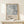 May include: Framed art poster with abstract blue and gray brushstrokes in wooden frame, leaning against white wall on light wood surface. Small wrapped white gift box with twine and greenery sits in front. Poster features exhibition-style text below the artwork. Soft natural lighting creates gentle shadows on the minimalist display.