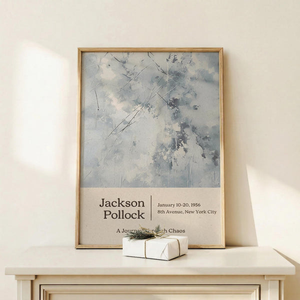 May include: Framed art poster with abstract blue and gray brushstrokes in wooden frame, leaning against white wall on light wood surface. Small wrapped white gift box with twine and greenery sits in front. Poster features exhibition-style text below the artwork. Soft natural lighting creates gentle shadows on the minimalist display.
