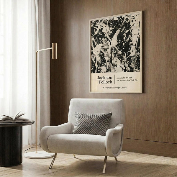 May include: Jackson Pollock exhibition poster with black and white abstract paint splatter artwork mounted on wood paneled wall above beige upholstered armchair with geometric patterned pillow. Modern reading corner features brass floor lamp with white circular base and dark round side table with open book. Natural lighting from window with white curtains illuminates warm wood grain textures and neutral color palette.
