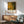 May include: Jackson Pollock exhibition poster with abstract yellow, green and brown organic patterns and black branching lines, displayed in wooden frame above mid-century modern credenza. Sage green upholstered chair with wooden arms sits nearby, white ceramic lamp on credenza surface, warm sunlight casting shadows across white wall in contemporary living room setting.