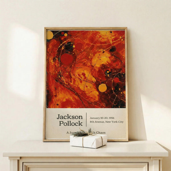 May include: Abstract expressionist artwork poster in wooden frame featuring bold red, orange, and black paint splatters with circular forms and dynamic brushstrokes. Museum-style exhibition poster with cream text area below the artwork. Displayed on light wood console table against white wall, with small wrapped gift box with twine and greenery sprig positioned in front.