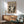 May include: Framed Jackson Pollock exhibition poster in warm wood frame hanging above mid-century modern wooden sideboard. Green upholstered armchair with wooden arms sits beside round coffee table. White ceramic table lamp on sideboard. Poster features black and white abstract expressionist artwork with dynamic paint splatters and gestural marks. Natural sunlight creates shadows across white wall and wooden furniture in contemporary living room setting.