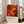 May include: Abstract expressionist art print in warm oranges, reds, and yellows with black paint splatters and drips, displayed in white frame leaning against bright white wall next to beige upholstered chair. Natural sunlight streams through tall windows creating soft shadows on light wooden floors with area rug.