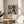 May include: Black and white abstract expressionist artwork in wooden frame displayed on round wooden side table next to beige upholstered chair. Sunlight streams across white wall creating geometric shadows. Green plant leaves visible in corner. Poster features dynamic paint splatters and drips with exhibition text below the artwork.