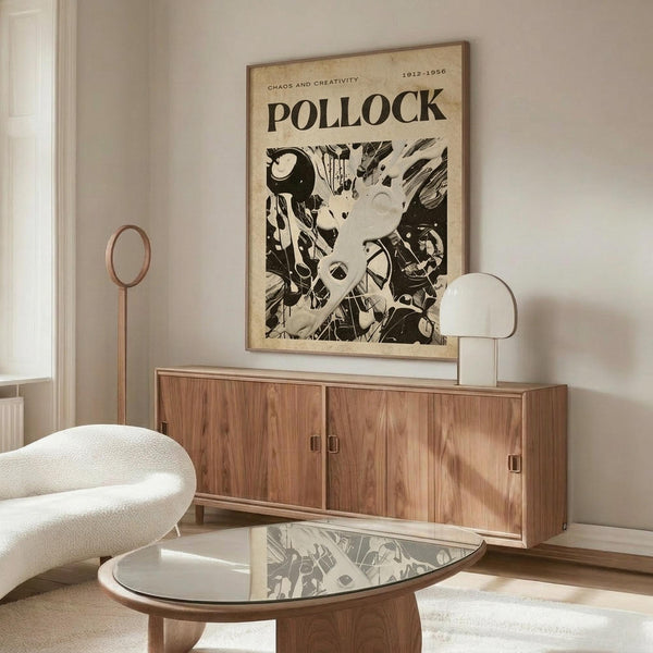 May include: Black and white Jackson Pollock art poster hanging above a mid-century modern walnut credenza in a minimalist living room. The vintage-style poster features abstract drip painting artwork with "POLLOCK" and "CHAOS AND CREATIVITY 1912-1956" text. Room has cream walls, white curved chair, oval glass coffee table, and warm wood furniture with clean lines.
