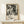 May include: Black and white abstract expressionist artwork in light wood frame, leaning against cream wall on pale wooden console table. Small white gift box with twine and evergreen sprig sits in front. Poster features chaotic paint splatters and drips with exhibition text below the artwork. Soft natural lighting creates gentle shadows on minimalist neutral-toned setting.