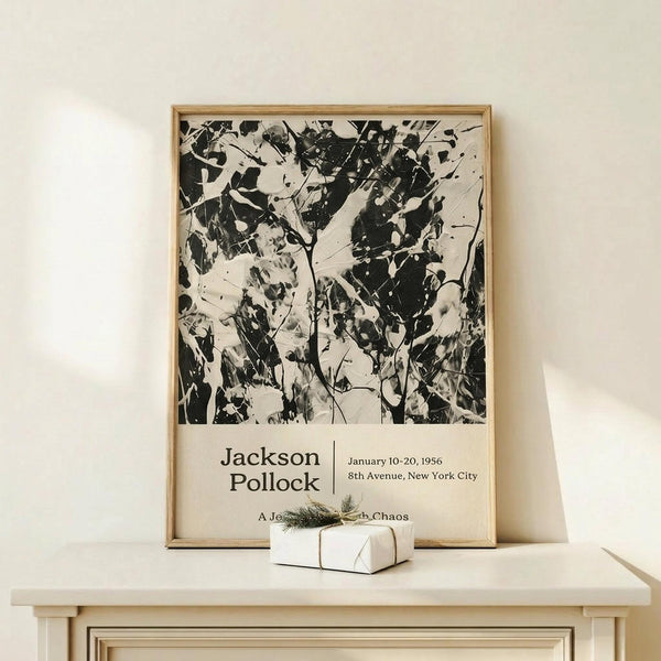 May include: Black and white abstract expressionist artwork in light wood frame, leaning against cream wall on pale wooden console table. Small white gift box with twine and evergreen sprig sits in front. Poster features chaotic paint splatters and drips with exhibition text below the artwork. Soft natural lighting creates gentle shadows on minimalist neutral-toned setting.
