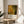 May include: Framed art print featuring Jackson Pollock's abstract expressionist style with organic yellow, green, and amber bubble-like forms connected by dark branching lines, displayed on a round wooden side table next to a beige upholstered chair with plant shadows cast on white walls in natural sunlight