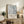 May include: Framed museum-style poster featuring abstract blue and gray paint splatters with diagonal lines, displayed on rustic wooden sideboard next to fern plant, books, ceramic vase and leather chair in neutral-toned living room setting