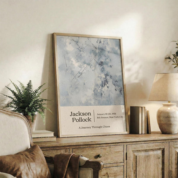 May include: Framed museum-style poster featuring abstract blue and gray paint splatters with diagonal lines, displayed on rustic wooden sideboard next to fern plant, books, ceramic vase and leather chair in neutral-toned living room setting