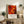 May include: Framed abstract art poster with vibrant red, orange and yellow swirling patterns and circular elements, displayed on rustic wooden console table alongside fern plant, ceramic vase, and stacked books in neutral-toned living room with beige armchair and cream walls