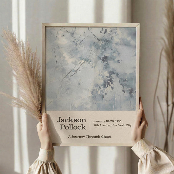 May include: Person holding framed abstract art poster with pale blue and gray brushstrokes and dark linear marks, featuring exhibition text for Jackson Pollock show from January 1956 in New York City with subtitle "A Journey Through Chaos." Dried pampas grass visible beside figure wearing cream ribbed sweater against soft neutral curtained background.
