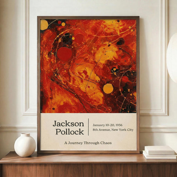 May include: Abstract expressionist poster featuring vibrant red, orange and yellow paint splatters with black curved lines and circular dots throughout. Exhibition-style design with artist name and show details in clean typography below the artwork. Framed print displayed on wooden sideboard alongside white ceramic vase and stacked books against cream paneled wall in modern interior setting.