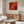 a modern living room with a large abstract painting hanging on the wall above a wooden cabinet. The painting has a vibrant mix of red, orange, and yellow colors, creating a striking visual contrast. The room also includes a white sofa, a glass coffee table, and a lamp on a side table.