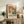 May include: Framed abstract art exhibition poster with orange, green and white paint textures displayed on rustic wooden sideboard next to fern plant, ceramic vase, and books in warm-toned living room setting with beige chair and lamp