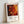 May include: Framed abstract art poster leaning against white wall, featuring vibrant red, orange, yellow and black fluid paint patterns with circular bubble-like forms. Light wood frame with white matting displays exhibition-style text below the artwork. Natural lighting creates soft shadows on the wall.