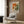 May include: Framed abstract art poster featuring Jackson Pollock-style paint splatters in orange, green, and cream colors with bold black text below. Displayed on wood-paneled wall above beige upholstered armchair with geometric patterned pillow. Modern brass floor lamp and black side table with open book complete the contemporary living room setting with natural lighting from nearby window.