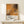 May include: Framed Jackson Pollock exhibition poster in natural wood frame, leaning against white wall on light wooden surface. Abstract orange and black paint splatter artwork dominates upper portion, with white text area below showing artist name and exhibition details. Small white wrapped gift with natural twine and evergreen sprig sits beside frame. Warm, minimalist styling with neutral tones throughout scene.