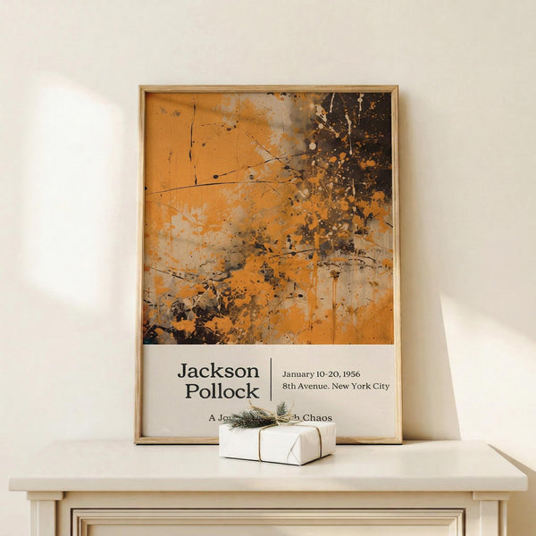 May include: Framed Jackson Pollock exhibition poster in natural wood frame, leaning against white wall on light wooden surface. Abstract orange and black paint splatter artwork dominates upper portion, with white text area below showing artist name and exhibition details. Small white wrapped gift with natural twine and evergreen sprig sits beside frame. Warm, minimalist styling with neutral tones throughout scene.