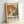 May include: Framed abstract art exhibition poster with orange, green and white paint splatters in Jackson Pollock style, displayed against white wall. Cream colored wooden frame contains vintage-style poster with "POLLOCK" text and exhibition dates "MAY 18, 2030 - AUGUST 14, 2030" beneath colorful abstract artwork resembling dripped paint technique.