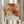 May include: Person in cream sweater holding framed abstract art poster featuring vibrant orange, red, and black paint splatters with white linear elements. Dried pampas grass visible to left. Poster shows exhibition text reading "Jackson Pollock January 10-20, 1956 8th Avenue, New York City" below the artwork. Light wooden frame against soft neutral curtained background.