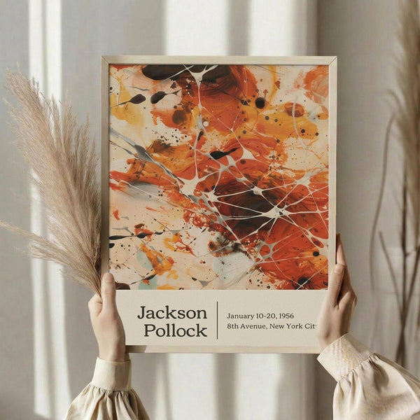 May include: Person in cream sweater holding framed abstract art poster featuring vibrant orange, red, and black paint splatters with white linear elements. Dried pampas grass visible to left. Poster shows exhibition text reading "Jackson Pollock January 10-20, 1956 8th Avenue, New York City" below the artwork. Light wooden frame against soft neutral curtained background.
