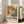 May include: Framed abstract art exhibition poster with orange, green and white paint splashes in Jackson Pollock style, displayed on light wooden floor against white wall corner. Gray upholstered sofa with pillows visible on right side. Natural sunlight streaming through tall windows creates soft shadows on wall. Poster features bold black text beneath colorful abstract artwork section.