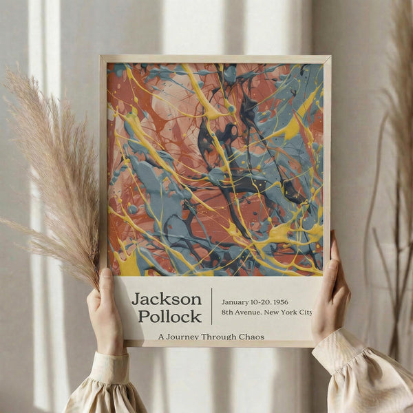 May include: Person holding framed abstract art poster featuring dynamic splashes and drips in orange, blue, and yellow colors reminiscent of Jackson Pollock's drip painting style. The poster shows exhibition details for a 1956 show at 8th Avenue, New York City with subtitle "A Journey Through Chaos." Light wood frame against neutral curtained background with dried pampas grass visible to the left.