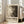 May include: Framed poster with abstract black and white splattered artwork leaning against white wall next to beige upholstered chair. Natural lighting from tall windows creates soft shadows on hardwood floor. The poster features chaotic paint splatters and drips transitioning from light cream on left to dark black on right, with exhibition-style text below reading "POLLOCK" and "EMBRACE THE CHAOTIC REALM OF CREATIVITY" with dates "MAY 18, 2030 - AUGUST 14, 2030".