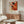 May include: Abstract expressionist poster with vibrant orange, red, and black paint splatters on cream background, featuring exhibition text below artwork. Displayed above mid-century modern walnut credenza in minimalist living room with white curved chair, glass oval coffee table, and brass floor lamp.