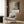 May include: A framed poster featuring abstract black and gray paint splatters on cream background with exhibition-style text below, displayed on wood-paneled wall above beige upholstered chair with geometric patterned pillow, next to brass floor lamp and dark round side table with open book.
