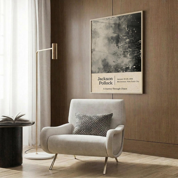 May include: A framed poster featuring abstract black and gray paint splatters on cream background with exhibition-style text below, displayed on wood-paneled wall above beige upholstered chair with geometric patterned pillow, next to brass floor lamp and dark round side table with open book.