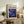 May include: Framed poster with cream background featuring "POLLOCK" text and "CHAOS AND CREATIVITY 1912-1956" header above swirling blue and purple abstract artwork with gold dots, displayed on rustic wooden sideboard alongside fern plant, ceramic vases, books, and brown leather chair in neutral-toned living room setting.