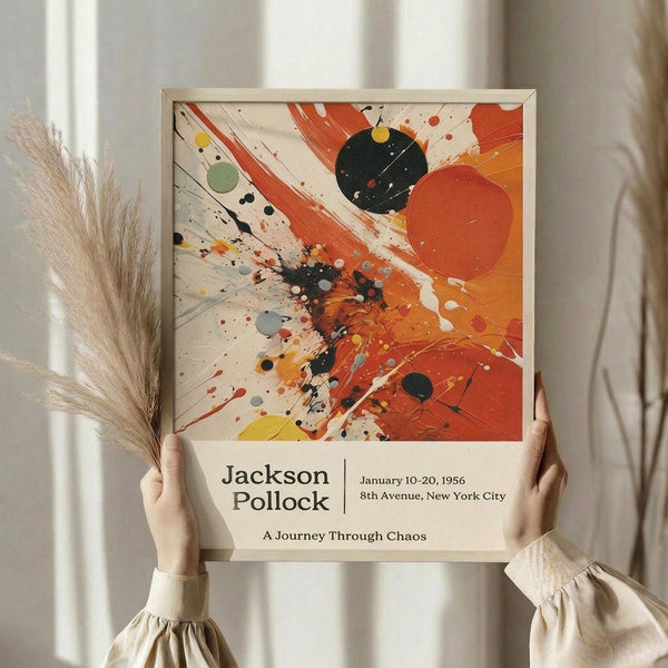 May include: Person holding framed poster featuring abstract expressionist artwork with vibrant orange, red, and black paint splatters and brushstrokes on cream background. Exhibition-style poster displays "Jackson Pollock" and "A Journey Through Chaos" text with vintage gallery details. Natural lighting highlights the dynamic paint texture and energetic composition while dried pampas grass appears in soft focus nearby.