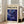 May include: Framed art poster with cream background featuring "POLLOCK" in bold black letters, "CHAOS AND CREATIVITY 1912-1956" text, and swirling blue abstract artwork with yellow and white dots. Natural wood frame leans against white wall next to beige upholstered chair in bright room with large windows and wooden floors.