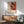 May include: A framed Jackson Pollock exhibition poster featuring abstract paint splatters in red, orange, black, and white with exhibition details. The poster hangs above a wooden credenza with a white lamp in a mid-century modern living room setting with a mint green armchair, round coffee table, and warm sunlight casting shadows on white walls.