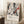 May include: Abstract expressionist artwork poster in light wood frame leaning against white wall. Features chaotic black paint splatters and drips with orange-red accents on cream background. Exhibition-style text below reads "Jackson Pollock" with dates and "A Journey Through Chaos." Scene shows cozy corner with white coffee cup on wooden block, cream knit blanket, and natural sunlight creating shadows on white walls and fluffy rug.