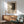 May include: Framed Jackson Pollock exhibition poster hanging above mid-century modern wooden credenza with white table lamp. Features abstract black and white splattered artwork with "POLLOCK" text and exhibition dates. Mint green upholstered armchair with wooden frame sits beside round coffee table. Warm sunlight creates geometric shadows on white wall in minimalist living room setting.
